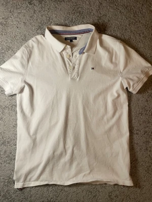 Vit pikétröja från Tommy Hilfiger - Klassisk vit pikétröja från Tommy Hilfiger med broderad logga på bröstet. Tröjan har korta ärmar, krage och knappar framtill. Tillverkad i mjuk bomull och har diskreta blå detaljer på insidan av kragen.