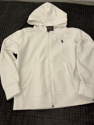Ralph lauren kofta  - Vit hoodie från Polo Ralph Lauren med klassisk svart logobroderi på bröstet. Tröjan har huva, dragkedja framtill och två fickor. Tillverkad i mjuk bomull, perfekt för chill dagar. Långärmad och med ribbade muddar för extra komfort.