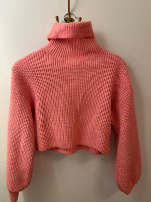 Croppad rosa polotröja stickad - Säljer en croppad stickad polotröja i en snygg rosa färg. Tröjan har lång ärm och en hög, bred polokrage som ger en cool vibe. Perfekt för dig som gillar trendiga och bekväma plagg med lite loose passform.