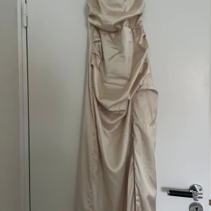 Så fin Champagnefärgad långklänning Bubbleroom - Supersnygg långklänning från Bubbleroom i champagnefärgat satinliknande material. Klänningen har figurnära passform, draperade detaljer i midjan och en hög slits framtill. Perfekt för dig som vill sticka ut med en elegant look.