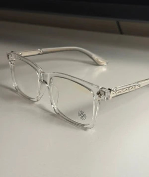 Transparenta glasögon från Chrome Hearts - Säljer ett par stilrena glasögon med transparent, rektangulär båge och coola silverdetaljer på skalmarna. Glasen har en liten blomma tryckt i hörnet för extra vibe. Perfekt för dig som vill sticka ut med en modern look. Dessa glaseögon är från märket  chrome hearts.