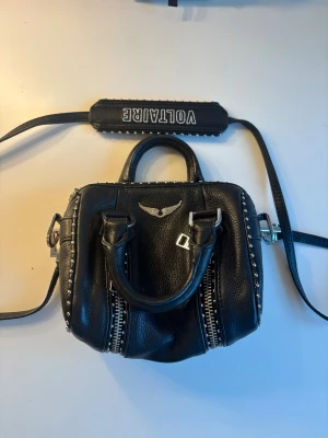 Svart handväska Zadig & Voltaire - Cool svart handväska från Zadig & Voltaire i skinn med silverfärgade nitar längs kanterna och detaljerade dragkedjor. Väskan har både handtag och axelrem med logotyp, samt ett snyggt emblem framtill. Perfekt storlek för det viktigaste och ger en edgy vibe.