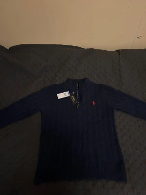Marinblå stickad tröja Polo Ralph Lauren - Snygg marinblå stickad tröja från Polo Ralph Lauren med half zip och klassisk kabelstickad design. Tröjan har långärmad passform, ribbad krage och röd broderad logga på bröstet. Perfekt för dig som gillar stilrena och tidlösa plagg.    Tröjan har storlek L men känns som medium därav säljer jag den.