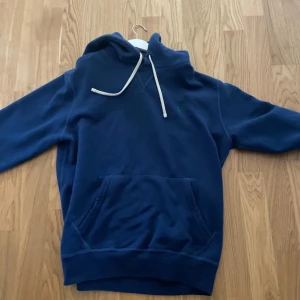 Marinblå hoodie från Polo Ralph Lauren - Snygg marinblå hoodie från Polo Ralph Lauren med klassisk huva och vita snören. Tröjan har en stor magficka framtill och diskret logga på bröstet. Perfekt för chill dagar och enkel att matcha med jeans eller joggers.