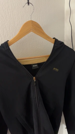 Svart kofta från Hugo Boss - Svart vindjacka från Hugo Boss med guldfärgad logga på bröstet och snygg guldfärgad dragkedja. Jackan har huva, diskreta fickor och en cool detalj med Boss-mönster längs ärmen. Perfekt för dig som gillar stilrena och sportiga plagg.