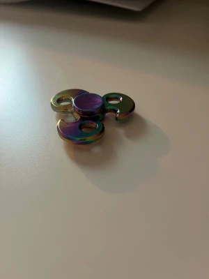 Metallisk Fidget Spinner - Snygg fidget spinner i metall med regnbågsfärgad finish. Trearmad design med rundade kanter och hål för extra grepp. Spinnern är i mycket gott skick utan synligt slitage. Perfekt för stresslindring eller som samlarobjekt.