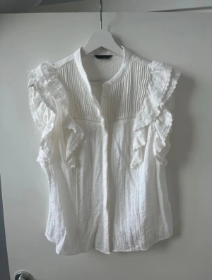 Vit blus med volang från Zara - Säljer en vit blus från Zara i storlek M. Blusen har volangdetaljer på axlarna, spetskant och knappar framtill. Materialet är lätt och luftigt, perfekt för varma dagar. Den har en klassisk krage och är ärmlös med snygga detaljer som ger en romantisk vibe.