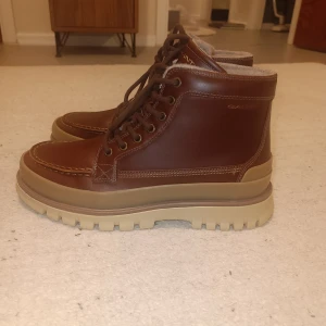 Bruna boots från Gant med snörning - Snygga bruna boots från Gant med grov beige sula och snörning framtill. Skorna är tillverkade i skinn och har rund tå samt kontrastsömmar. Insidan är fodrad med mjukt material för extra komfort. Perfekta för höst och vinter när du vill ha både stil och funktion.