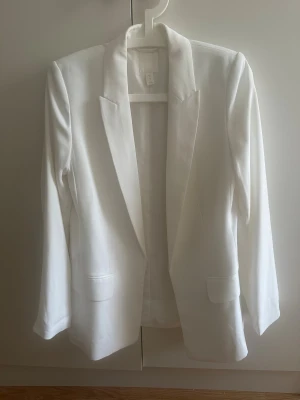 Vit kavaj från H&M - Snygg vit kavaj från H&M med klassisk krage och två fickor framtill. Rak passform och stilren design som ger en fräsch look. Matrialet är mjukt och lätt, perfekt för att lyfta din outfit. Passar dig som gillar minimalistisk stil och ha något som sticker ut