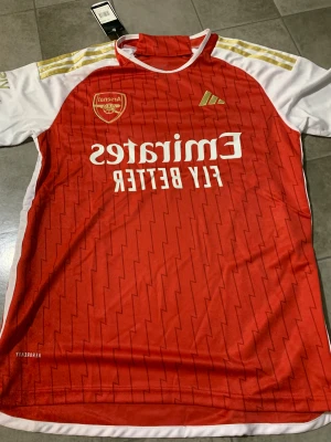 Arsenal hemmatröja Adidas S - Arsenal hemmatröja från Adidas i rött och vitt med guldiga detaljer och blixtmönster. Klassisk klubbsköld, Emirates-sponsor och tre guldiga ränder på axlarna. Tillverkad i lätt och ventilerande polyester, perfekt för match eller träning.