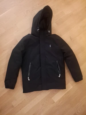  Polo Ralph Lauren Jacka - Svart vinterjacka från Polo Ralph Lauren med huva och två snedställda fickor med dragkedja. Jackan har det klassiska Polo-logot broderat på bröstet och elastiska muddar vid ärmsluten. Perfekt för dig som gillar stilren och sportig look. PRIS KAN DISKUTERAS