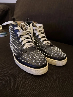 Christian Louboutin svarta sneakers med nitar - Svarta high-top sneakers från Christian Louboutin med vita snören och ikonisk röd sula. Skorna är täckta med silverfärgade nitar över hela ovandelen och har en platt vit sula. Tillverkade i skinn med lyxig känsla och edgy design som sticker ut.