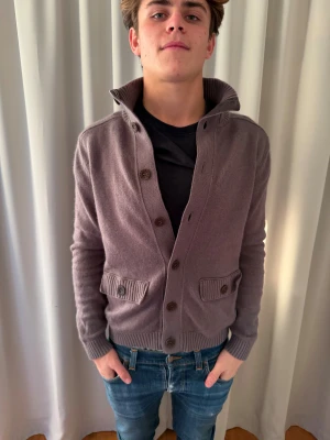 Brun stickad kofta från Strellson - Säljer en brun stickad kofta från Strellson i storlek S. Koftan har ribbad krage, knappstängning framtill och två fickor med knappdetalj. Materialet är mjukt och skönt, perfekt för lager-på-lager. Passformen är normal och ärmarna är långa.