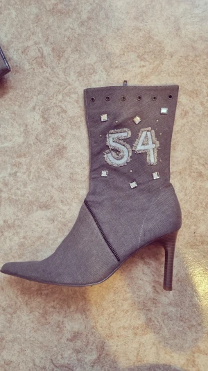 Grå stövletter med spetsig tå och klack - Unika grå stövletter med spetsig tå och hög klack. Dekorerade med broderade siffror '54', silvriga nitar och fyrkantiga paljetter. Materialet är manchester och skon har dragkedja på sidan för enkel påtagning. Perfekt för dig som gillar statement-detaljer.