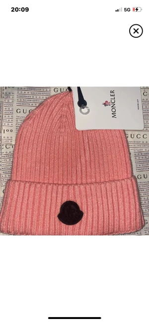 Rosa ribbad mössa från Moncler - Säljer en snygg rosa ribbstickad mössa från Moncler med uppvikt kant och svart logotyp framtill. Perfekt accessoar för att hålla värmen och ge din outfit en cool touch. Klassisk passform och mjukt material.