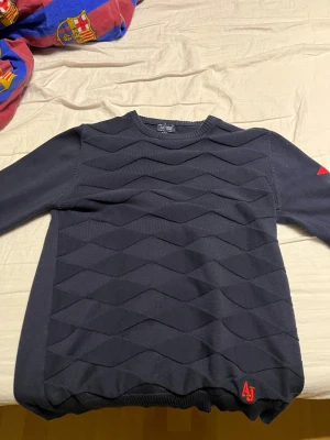 Mörkblå stickad tröja Armani Jeans - Snygg mörkblå stickad tröja från Armani Jeans med coolt geometriskt mönster på framsidan. Röd AJ-logga nertill och röd detalj med örn på ena ärmen. Rund hals och långa ärmar, perfekt för dig som gillar stilrena men unika plagg.PRIS KAN DISKUTERAS!