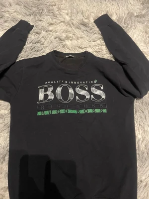 Svart sweatshirt från Hugo Boss M - Svart sweatshirt från Hugo Boss i storlek M med stort BOSS-tryck i vitt och grönt på bröstet. Tröjan har rund hals, långa ärmar och ribbade muddar. Materialet är mjuk bomull och passformen är normal. Perfekt för dig som gillar stilrena logotröjor.