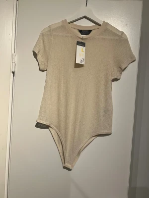 Beige ribbad bodysuit från Primark - Säljer en beige bodysuit från Primark i storlek L. Den har korta ärmar, rund hals och är ribbad i materialet. Bodyn är tunn och lätt transparent, perfekt för lager-på-lager. Enkel och clean design som funkar till många olika stilar.