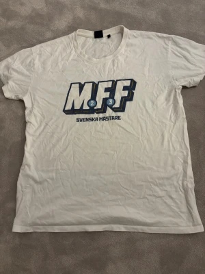 MFF t-shirt - Säljer en vit t-shirt med MFF-tryck och texten 'Svenska Mästare' på bröstet, i bra kvalitet. Materialet är mjuk bomull och trycket har blå och gula detaljer.