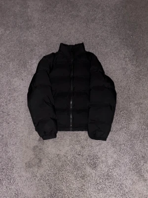 Svart pufferjacka från Weekday - Svart pufferjacka från Weekday i storlek S. Jackan har hög krage, lång ärm och dragkedja framtill. Den är quiltad och har en loose passform som ger en chill vibe. Perfekt för kalla dagar när du vill hålla dig varm och se cool ut.