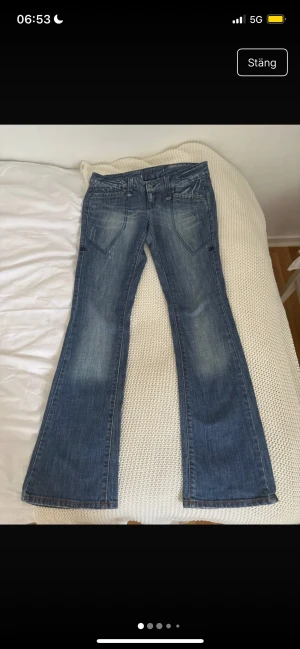 Blå bootcut jeans från Aino - Snygga blå jeans från Aino med bootcut passform och coola slitningar. Jeansen har stora framfickor med markerade sömmar och klassiska bakfickor med bruna detaljer. Materialet är jeans och midjan är normal. Perfekt för dig som gillar retrovibbar och breda ben.