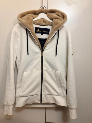 Moose Knuckles Bunny - Snygg vit jacka från Moose Knuckles. Köpt i 2024. Gold series. Kvitto och alla Tags finns med  den har mjuk beige fuskpäls i luvan, guldfärgade detaljer på ärmen och svart snörning. Jackan har dragkedja framtill, två fickor och ribbade muddar. Perfekt för dig som gillar streetwear med lyxig touch.