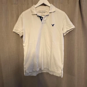 Vit pikétröja från American Eagle - Vit pikétröja från American Eagle Outfitters med klassisk krage och korta ärmar. Tröjan har en liten mörkblå logga broderad på bröstet och knäppning vid halsen. Tillverkad i mjuk bomull för en skön känsla.