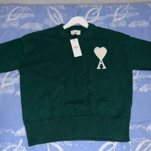 Grön stickad tröja från Ami Paris - Mörkgrön stickad tröja från Ami Paris med rund hals och ribbade muddar. På bröstet finns ett vitt hjärta med bokstaven A. Perfekt för dig som gillar stilrena och trendiga plagg med en twist.