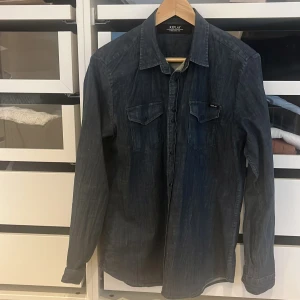 Mörkblå jeansskjorta från Replay - Snygg mörkblå jeansskjorta från Replay med klassisk krage och två bröstfickor med knappar. Skjortan har långa ärmar och en cool tvättad denimlook. Perfekt för dig som gillar streetstyle och vill ha en tidlös vibe i garderoben.
