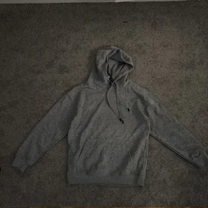 Grå hoodie från Polo Ralph Lauren - Snygg grå hoodie från Polo Ralph Lauren i storlek M. Tröjan har huva med snörning, ribbade muddar och klassisk broderad logga på bröstet. Perfekt för chill dagar och enkel att matcha med jeans.