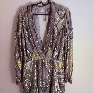 Glittrig silver kortklänning från ASOS - Säljer en glittrig kortklänning från ASOS i silver med paljetter och pärldetaljer. Klänningen har lång ärm, djup v-ringning och geometriskt mönster som verkligen sticker ut. Perfekt för dig som vill synas och älskar att glittra! Materialet är polyester.