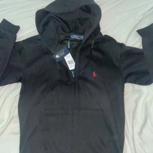 Svart hoodie från Polo Ralph Lauren - Svart hoodie från Polo Ralph Lauren med dragkedja och huva. Klassisk röd logga broderad på bröstet och snörning i huvan. Tillverkad i mjuk bomullsmix, perfekt för chill dagar. Fickor framtill för extra casual vibe.