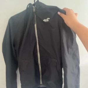 Svart vindjacka från Hollister - Svart vindjacka från Hollister med vit logga på bröstet och hel dragkedja framtill. Jackan har huva med snörning och två fickor. Materialet är lätt och syntetiskt, perfekt för blåsiga dagar. Stilren och enkel design som funkar till många outfits.
