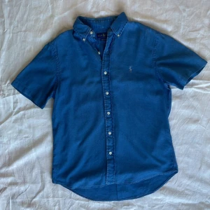 Blå kortärmad skjorta från Ralph Lauren - Snygg blå skjorta från Ralph Lauren med korta ärmar och klassisk button-down krage. Skjortan har vita knappar framtill och den ikoniska broderade loggan på bröstet. Tillverkad i mjuk bomull, perfekt för en chill och stilren look.
