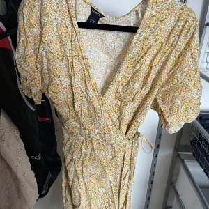 Blommig omlottklänning från Lindex - Säljer en söt omlottklänning från Lindex med blommigt mönster i gult, grönt och rosa på vit botten. Klänningen har korta puffärmar, knyt i midjan och v-ringad hals. Perfekt för sommarens alla tillfällen!