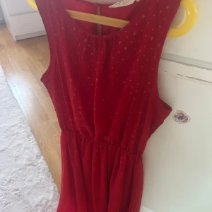 Röd kortklänning med stjärnmönster - Snygg röd kortklänning från H&M med silverfärgade stjärnor över hela tyget. Klänningen är ärmlös, har rund hals och ett litet knappslå i nacken. Midjan är lätt rynkad för en skön passform. Perfekt för dig som gillar färg och coola detaljer!