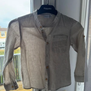 Randig henley-skjorta från Name It - Snygg henley-skjorta från Name It i ljusbeige med tunna ränder. Skjortan har lång ärm, träknappar framtill och en bröstficka. Tillverkad i mjuk bomull som känns skön mot huden. Perfekt för dig som gillar enkel och stilren design.