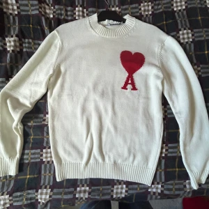 Vit stickad tröja från Ami Paris - Vit stickad tröja från Ami Paris med röd hjärtlogga och bokstaven A på bröstet. Klassisk rund halsringning och ribbade muddar vid ärmslut och nederkant. Helt oanvänd så den är i nytt skick  priset är inte hugget i sten. Säljer för den inte passar.