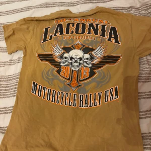 Affliction type motorcykel t shirt  - Cool brun t-shirt från Laconia Motorcycle Rally USA 2013 med stort tryck på ryggen med döskallar, vingar och orange detaljer. Framsidan har mindre tryck med samma motiv. Klassisk passform och rund hals, perfekt för dig som gillar bikerstil.