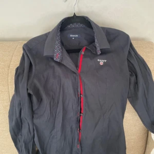 Mörkblå skjorta från GANT med röd detalj - Snygg mörkblå skjorta från GANT med coola röda detaljer längs knappraden och broderad logga på bröstet. Kragen har ett prickigt mönster på insidan som ger extra stilpoäng. Skjortan har långa ärmar och klassisk passform, perfekt för dig som gillar att sticka ut lite.