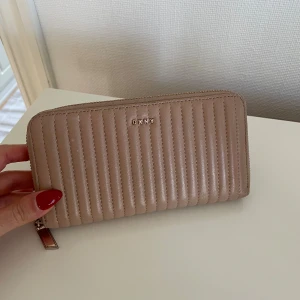 Beige quiltad plånbok från DKNY - Snygg beige plånbok från DKNY i quiltad design med vertikala sömmar. Plånboken är tillverkad i skinn och har en dragkedja runtom samt guldfärgad logga framtill. Perfekt storlek för att få plats med både kort och kontanter.