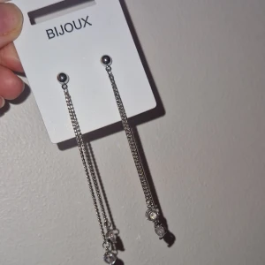 Långa silverfärgade örhängen Bijoux - Snygga nya långa örhängen från Bijoux i silverfärg med tunna kedjor och glittrande stenar längst ner. Perfekta för att ge en extra touch till din look. Örhängena har en minimalistisk design med små runda detaljer och gnistrande stenar som fångar ljuset.