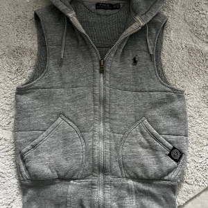 Grå hoodie-väst från Polo Ralph Lauren - Grå väst med huva från Polo Ralph Lauren. Västen har dragkedja framtill, två stora fickor och snörning i huvan. Tillverkad i mjuk bomullsmix och har det klassiska Polo-broderiet på bröstet. Perfekt för lager-på-lager och chill stil.