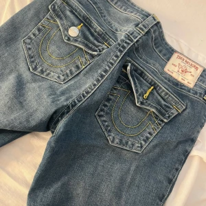 True Religion blå bootcut jeans - Snygga blå jeans från True Religion med klassiska gula sömmar och ikoniska ficklock bak. Modellen har bootcut-ben och silverfärgade knappar. Jeansen är tillverkade i mjukt denim och har en normal passform med medelhög midja.