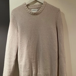 Beige stickad tröja från East West - En stilren beige stickad tröja från East West med rund hals och ribbade detaljer vid halsringning och nederkant. Tröjan har långärmad passform och är perfekt för dig som gillar enkel och clean look. Materialet är mjukt och känns skönt mot huden.