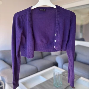 Lila croppad kofta med glittriga knappar - Superfin lila croppad kofta från H&M med långa ärmar och tre glittriga knappar framtill. Koftan har ribbade muddar och en mjuk, stickad känsla. Perfekt att slänga över en topp för en cool och färgstark look.