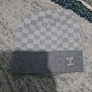 Grå rutig mössa från L&V - Snygg grå mössa från L&Vmed klassiskt rutigt mönster och vikt kant. Mössan har en broderad L&V-logga framtill och är stickad i mjukt material. Perfekt accessoar för att lyfta din vinterstil.