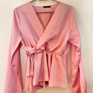 Rosa omlottblus från Missay - Säljer en söt rosa omlottblus från Missay med knyt i midjan och v-ringad hals. Blusen har långa ärmar och ett lätt glansigt tyg som ger en lyxig känsla. Perfekt för dig som gillar feminina detaljer och en snygg siluett.