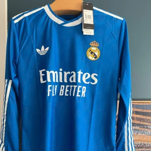 Blå Real Madrid långärmad fotbollströja - Snygg blå långärmad Real Madrid fotbollströja från Adidas med vita detaljer och klubbmärke på bröstet. Tröjan har vit krage, vita ränder längs ärmarna och subtilt mönster i tyget. Materialet är lätt och andas, perfekt för träning eller match.
