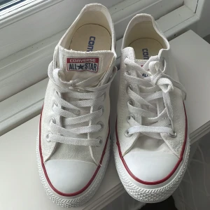 Vita Converse All Star sneakers, strl 38 - Säljer ett par klassiska vita Converse All Star sneakers i storlek 38. Skorna har rund tå, platt sula och snörning. De är låga med röd och blå rand längs sulan, tillverkade i textil med gummisula. Perfekta för dig som gillar tidlös streetstil.